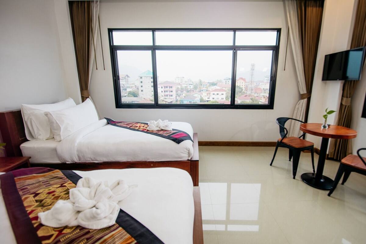 A hotel room with a bed, a small table, two chairs, a TV, and a large window with a city view.