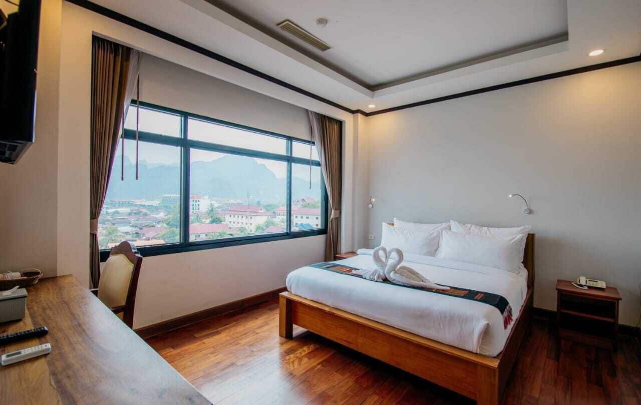 Una habitación de hotel con un ventanal grande, una cama de madera, una mesita de noche y un televisor.