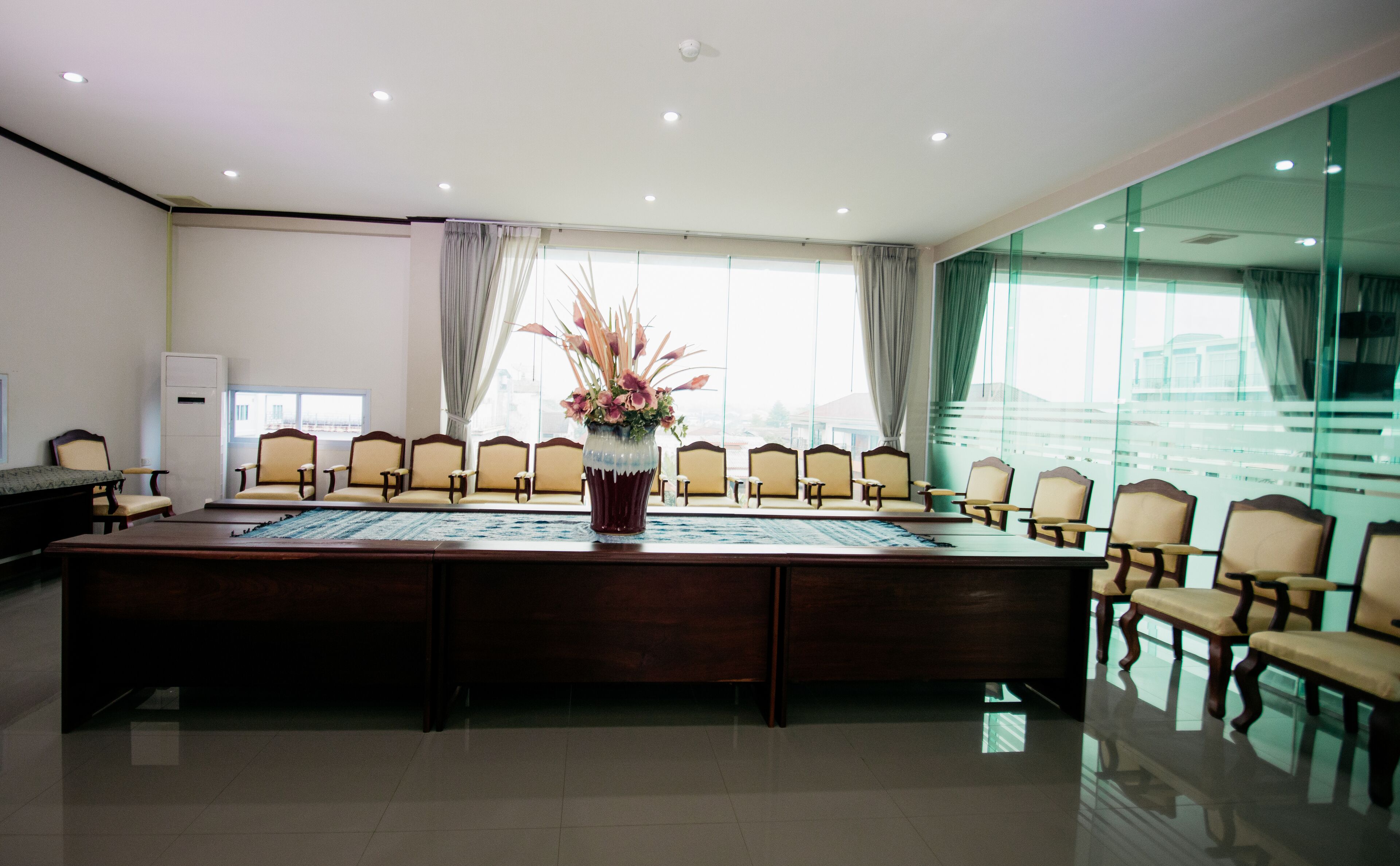 Un salón de conferencias con una larga mesa, varias sillas, un florero y ventanas amplias con cortinas.
