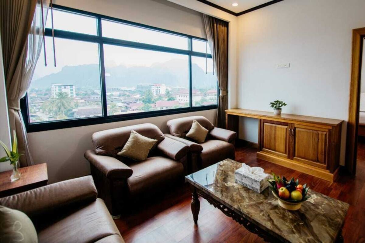 A living room with a sofa set, a coffee table with fruit, and a view of a cityscape through large windows.
