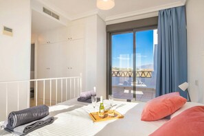 A hotel room with a bed, a small table with a bottle and glasses, a balcony with a view, and a window with curtains.