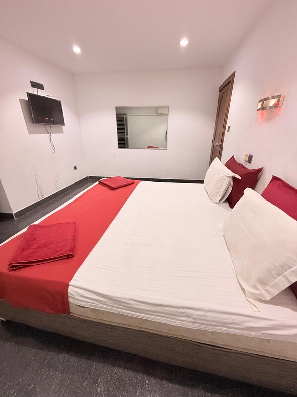 A hotel room with a large bed, red and white bedding, a flat-screen TV mounted on the wall, a small window, and a door.