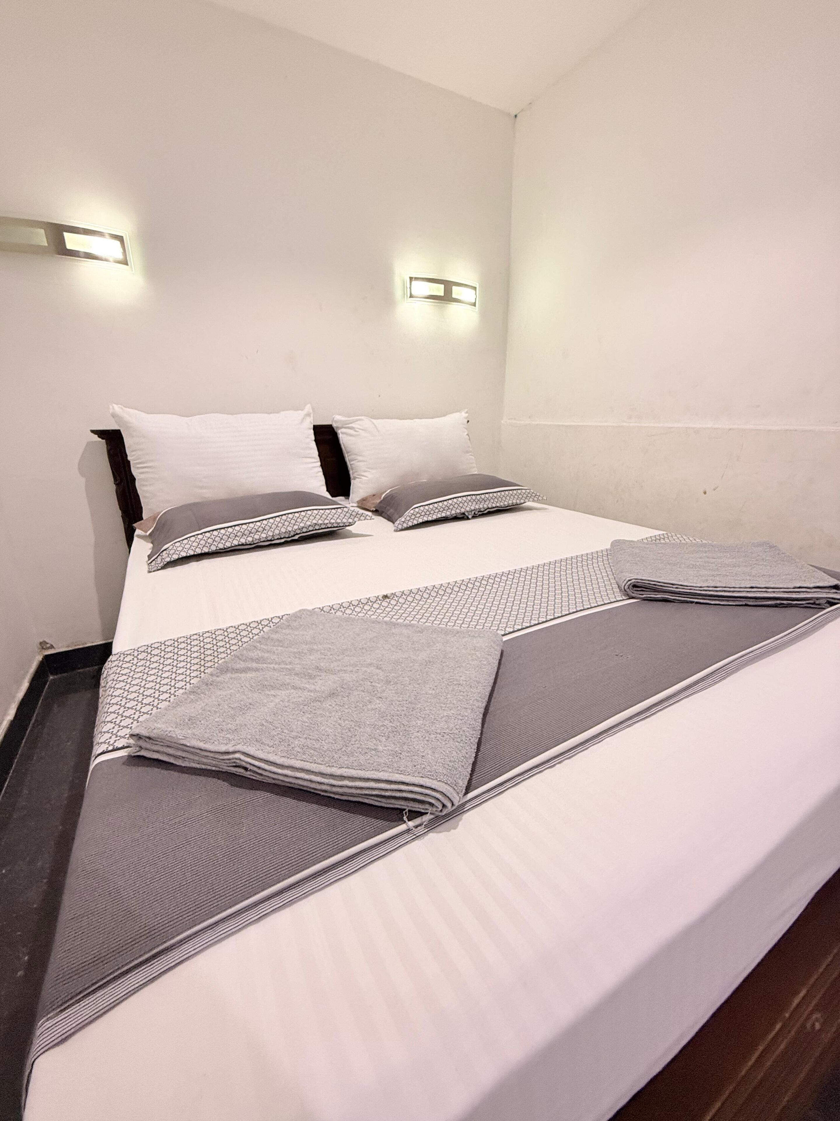 A neatly made bed with white linens and gray blankets, two wall-mounted light fixtures, and a plain white wall.