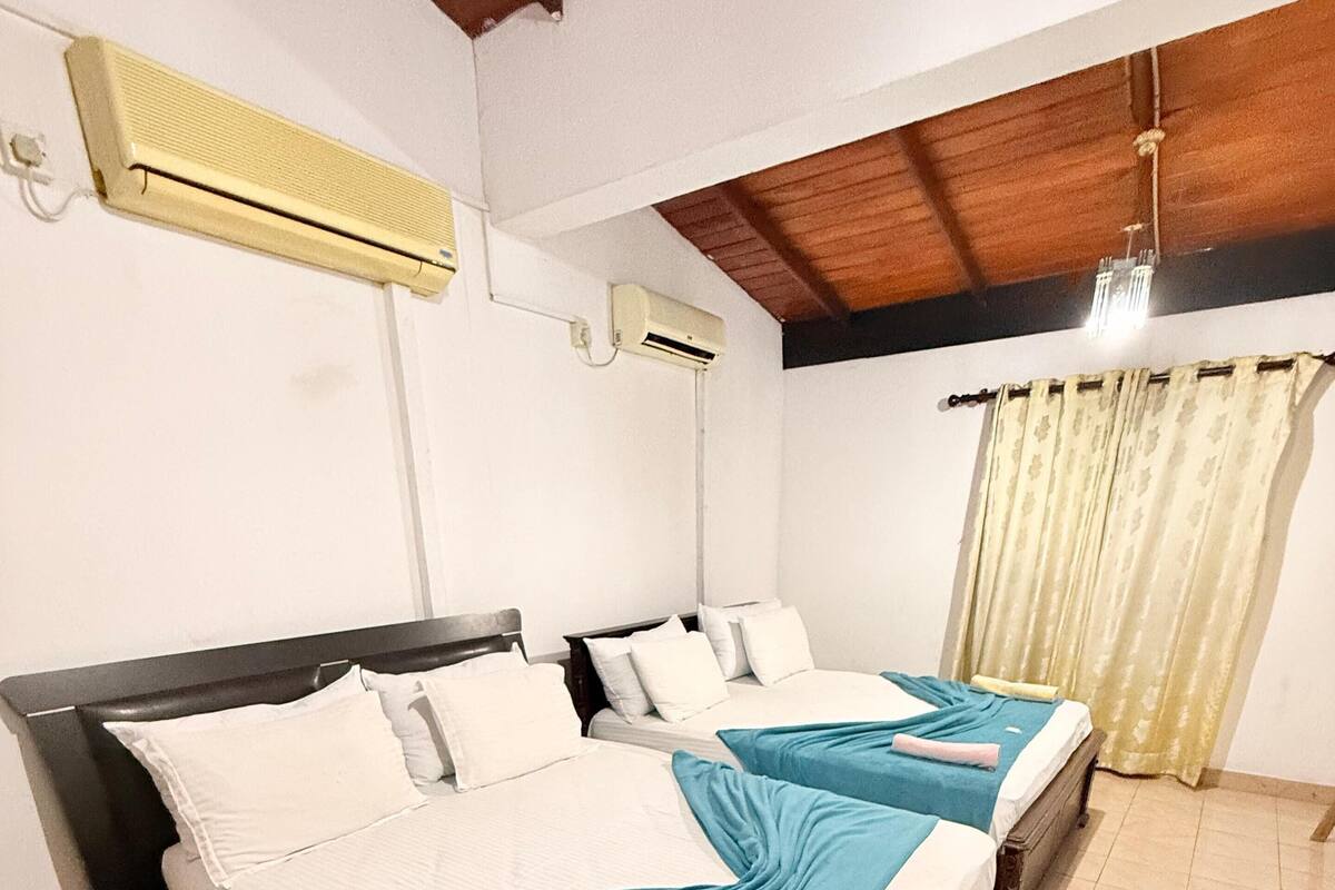 A room with two beds, each with white linens and teal bedspreads. There are ceiling fans and an air conditioning unit on the walls.