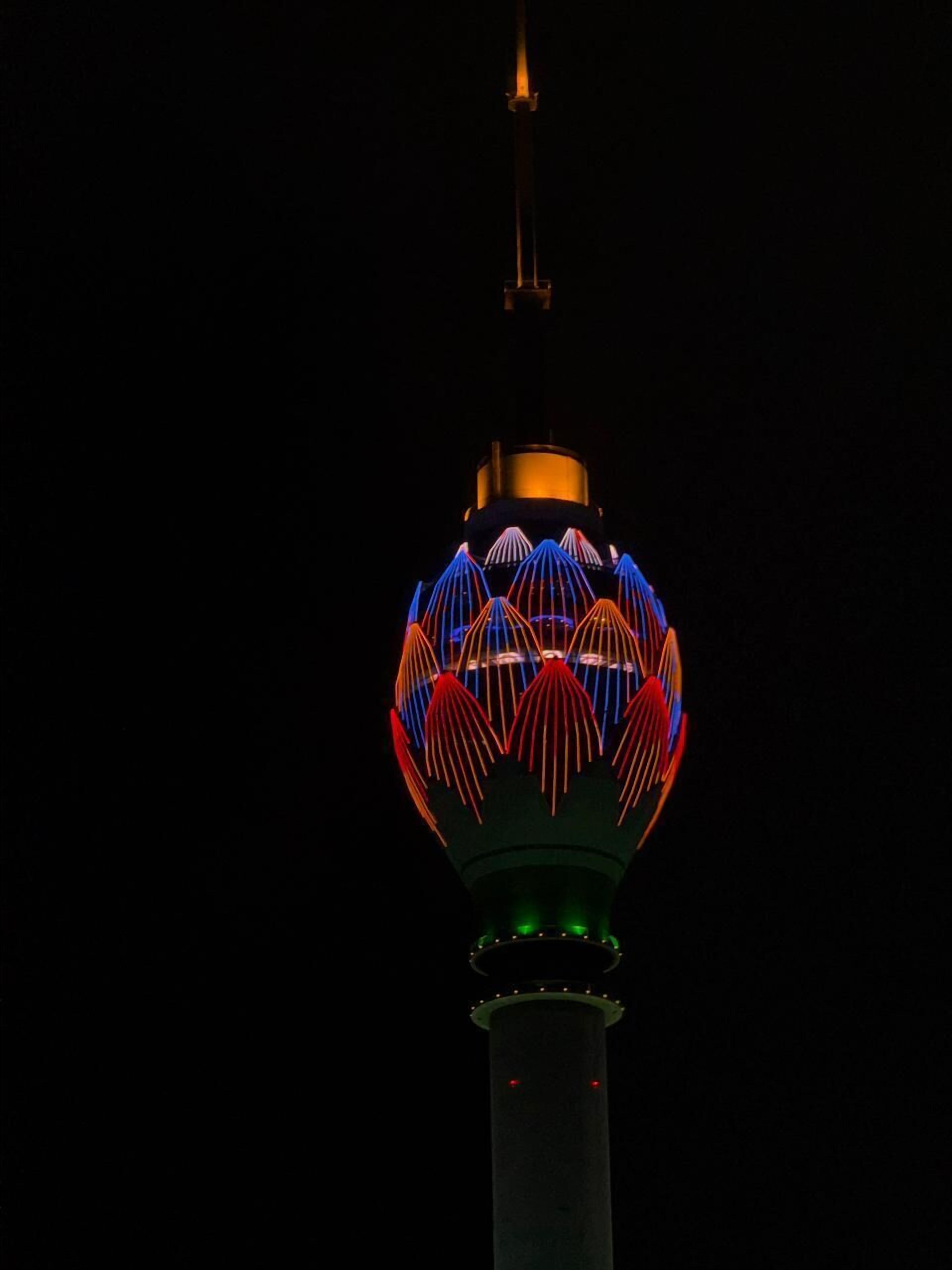 A tower with a spherical structure at the top, featuring a pattern of lines and illuminated in red and blue lights against a dark sky.