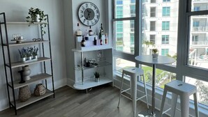A modern living room with a wooden floor, a white shelving unit with plants, a round table with two stools, and a large window with a view of buildings. - View with a View Waterfront WiFi,Furnished,Beaches,New Modern (Tampa)