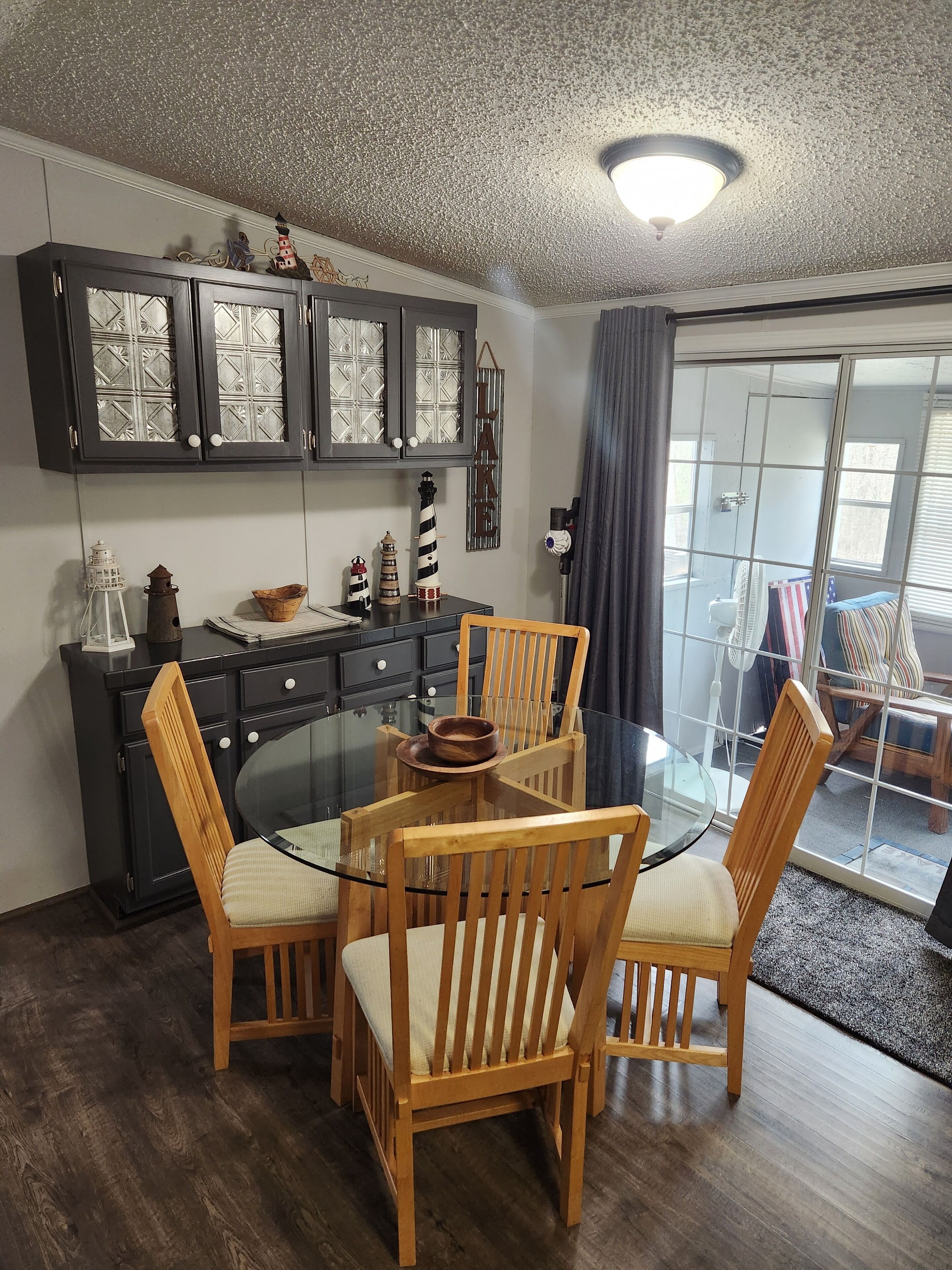Un coin repas avec une table en verre entourée de chaises en bois, un buffet en bois foncé doté de portes en verre, et une vue sur un balcon, accessible par une porte en verre.