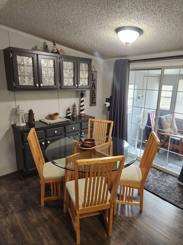 A dining area with a glass table surrounded by wooden chairs, a dark wooden cabinet with glass doors, and a view through a glass door to a balcony.