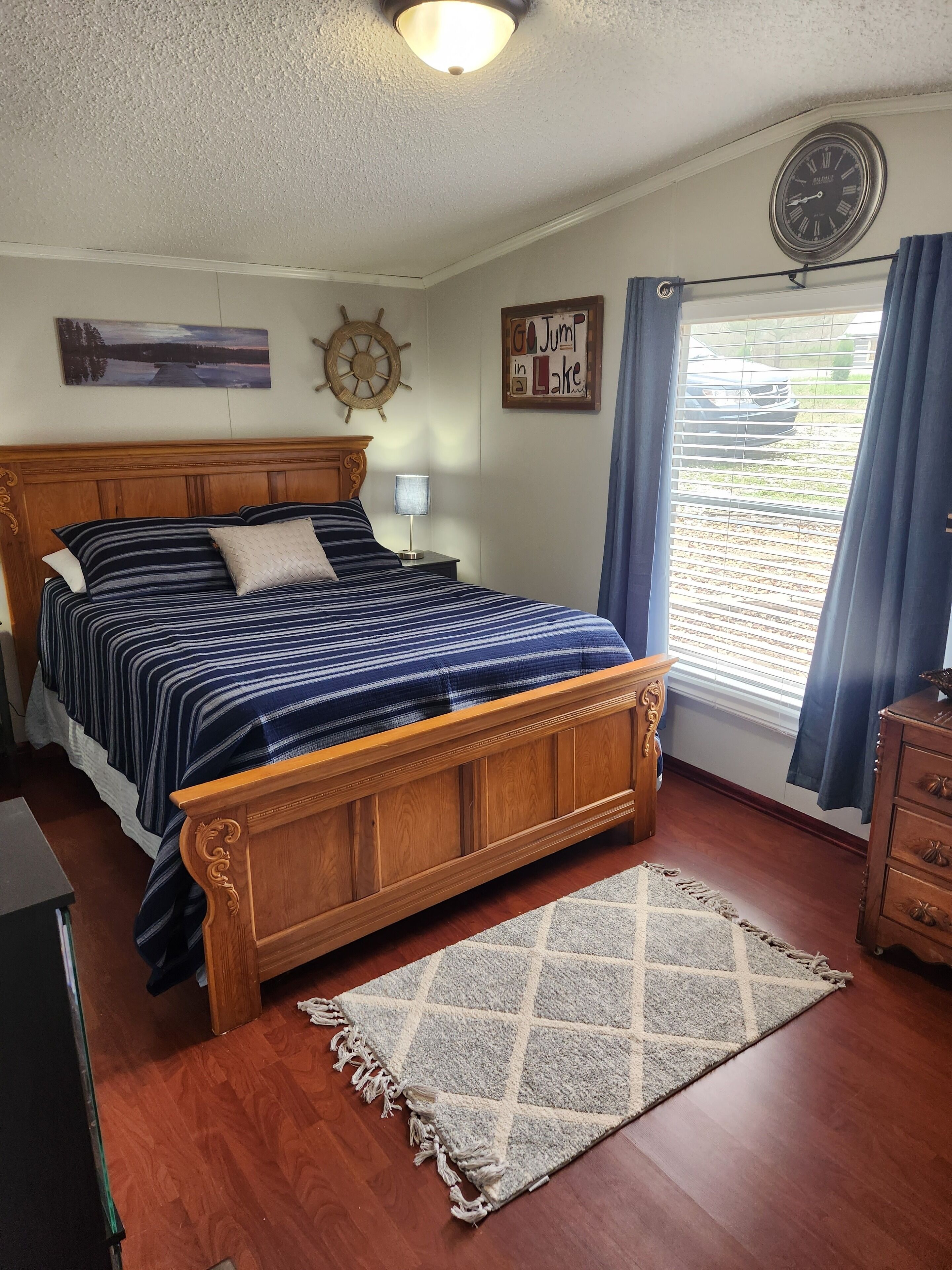 Une chambre à coucher avec un lit en bois, une horloge murale, un décor représentant un gouvernail de bateau, un tableau encadré et une fenêtre avec des rideaux.