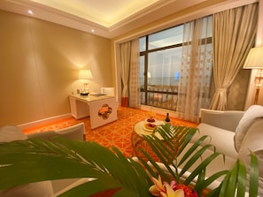 A hotel room with a large window, a desk with a lamp, a sofa, and a dining table with a cake and flowers. - Ramada by Wyndham Sihanoukville Xinhaohui Palace (Sihanoukville)
