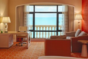 Habitación de hotel con escritorio, una silla, un sofá, una mesita y vistas al mar a través de amplias ventanas.
