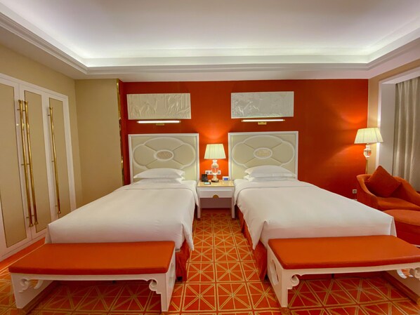 A hotel room with two beds, a nightstand with lamps, a bench, and a patterned floor.