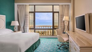 Una camera d'albergo con un letto, una televisione, una scrivania, una sedia e vista mare attraverso un'ampia finestra.
