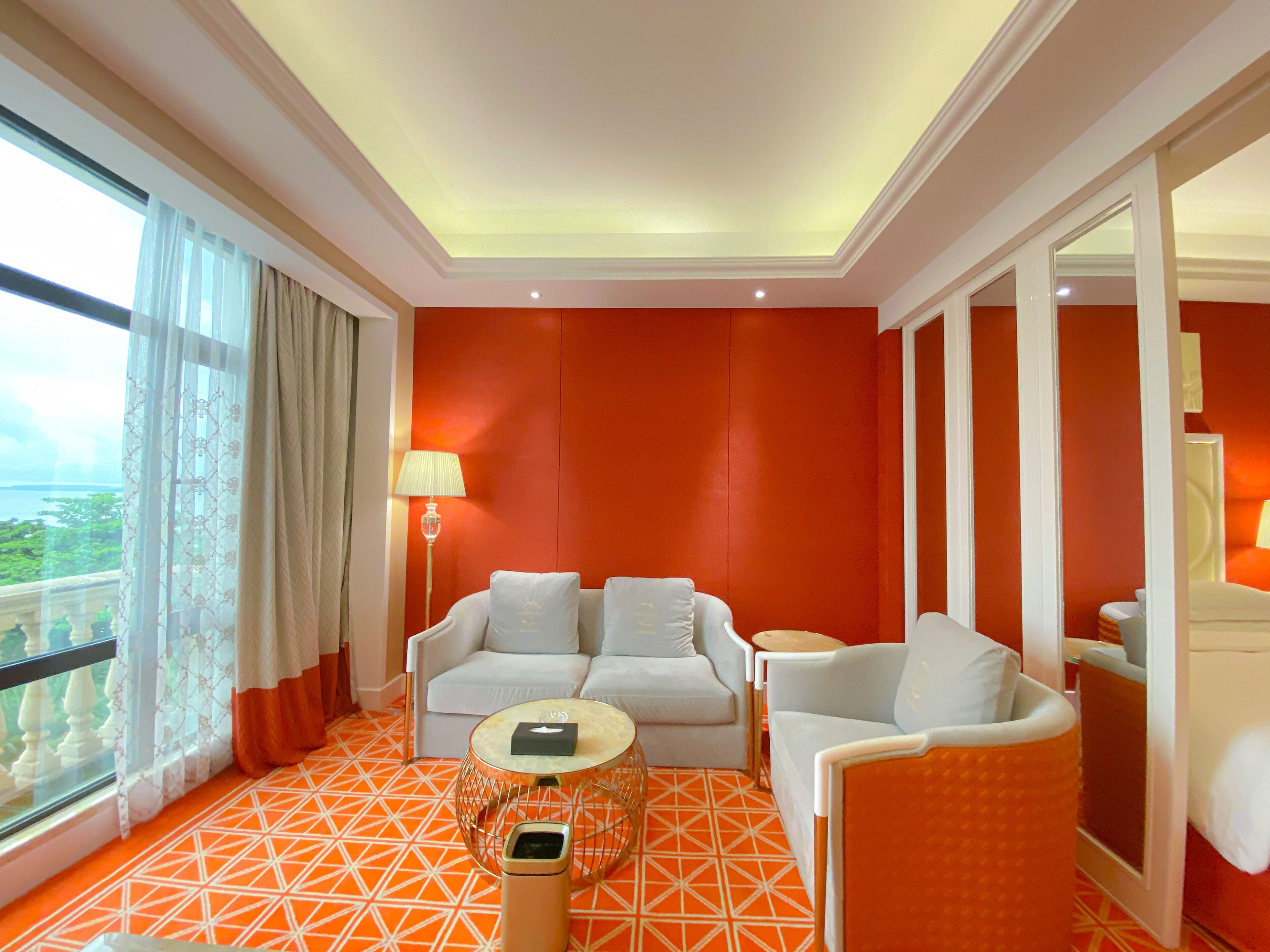 A hotel room with a red accent wall, a white sofa, a white armchair, a small round table, and a floor lamp.