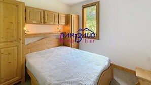 Une chambre à coucher avec des meubles en bois, un lit recouvert d’une courtepointe blanche à motifs, et une fenêtre donnant sur un paysage verdoyant.