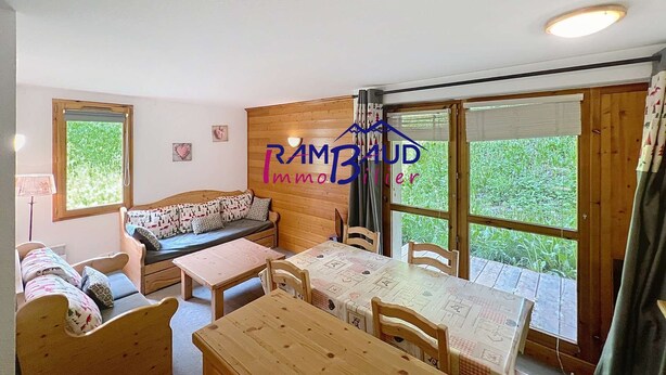 Une pièce chaleureuse avec un canapé en bois, une petite table et une table à manger. Il y a deux fenêtres avec des rideaux, et un mur en bois orné d’un logo.