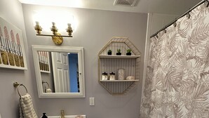 A bathroom with a white sink, a toilet, a shower curtain with a leaf pattern, a mirror, and a shelf with plants and jars. - Hamptons Apartment (East Quogue)