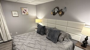 A bedroom with a large bed, a nightstand, a wall clock, and a decorative wall piece. - Hamptons Apartment (East Quogue)