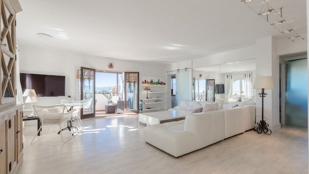 A spacious living room with a large white sofa, a white coffee table, and a white chair. There is a flat-screen TV mounted on the wall, a lamp on a side table, and a large mirror reflecting the room. - Spectacular apartment in Mont-roig (Mont-roig del Camp)