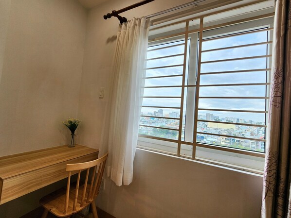 Una habitación con un escritorio de madera, una silla, un ventanal con vistas a un paisaje urbano y una cortina.