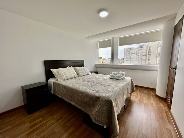 A bedroom with a bed, a nightstand, a window with a city view, and a door.