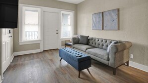 Una sala de estar con un sofá gris, un otomano azul y dos ventanas con persianas.