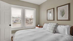 Un dormitorio con una cama grande, dos ventanas con persianas, una mesita de noche con un reloj y dos láminas enmarcadas con motivos botánicos en la pared.