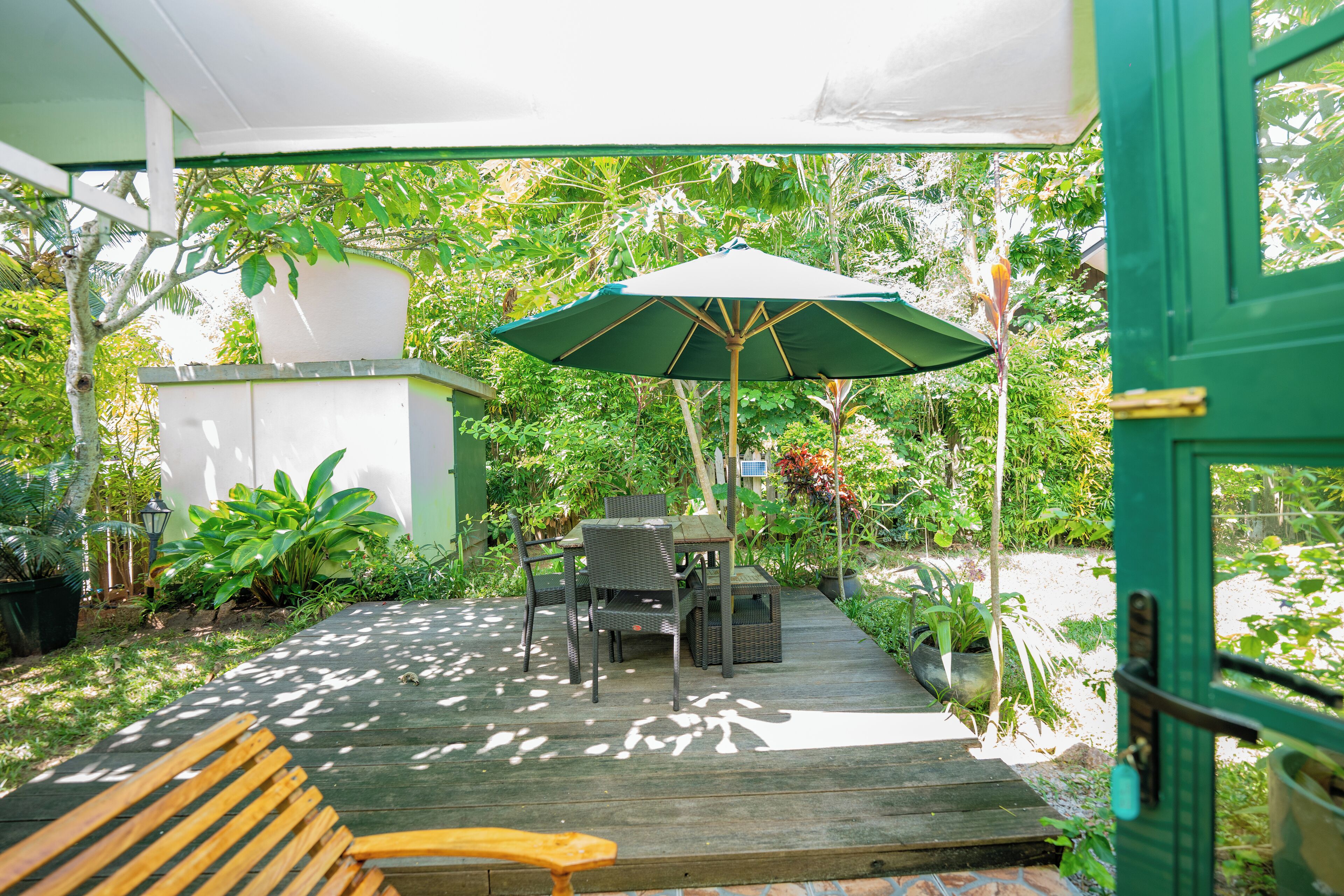 Un patio exterior con una mesa y sillas de madera, una sombrilla verde y plantas en macetas.