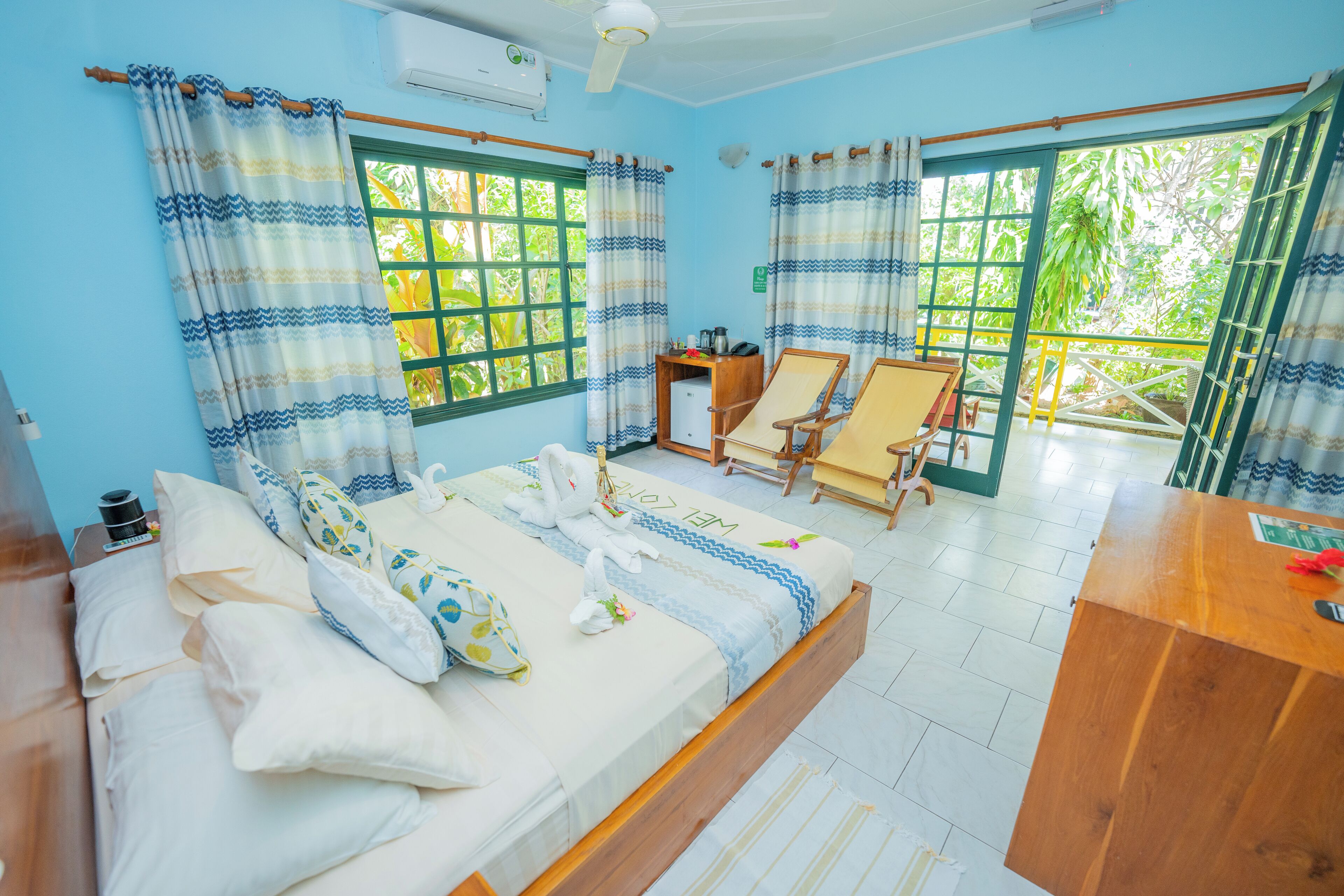 A room with a bed, a desk, two chairs, and a view of greenery through the windows.