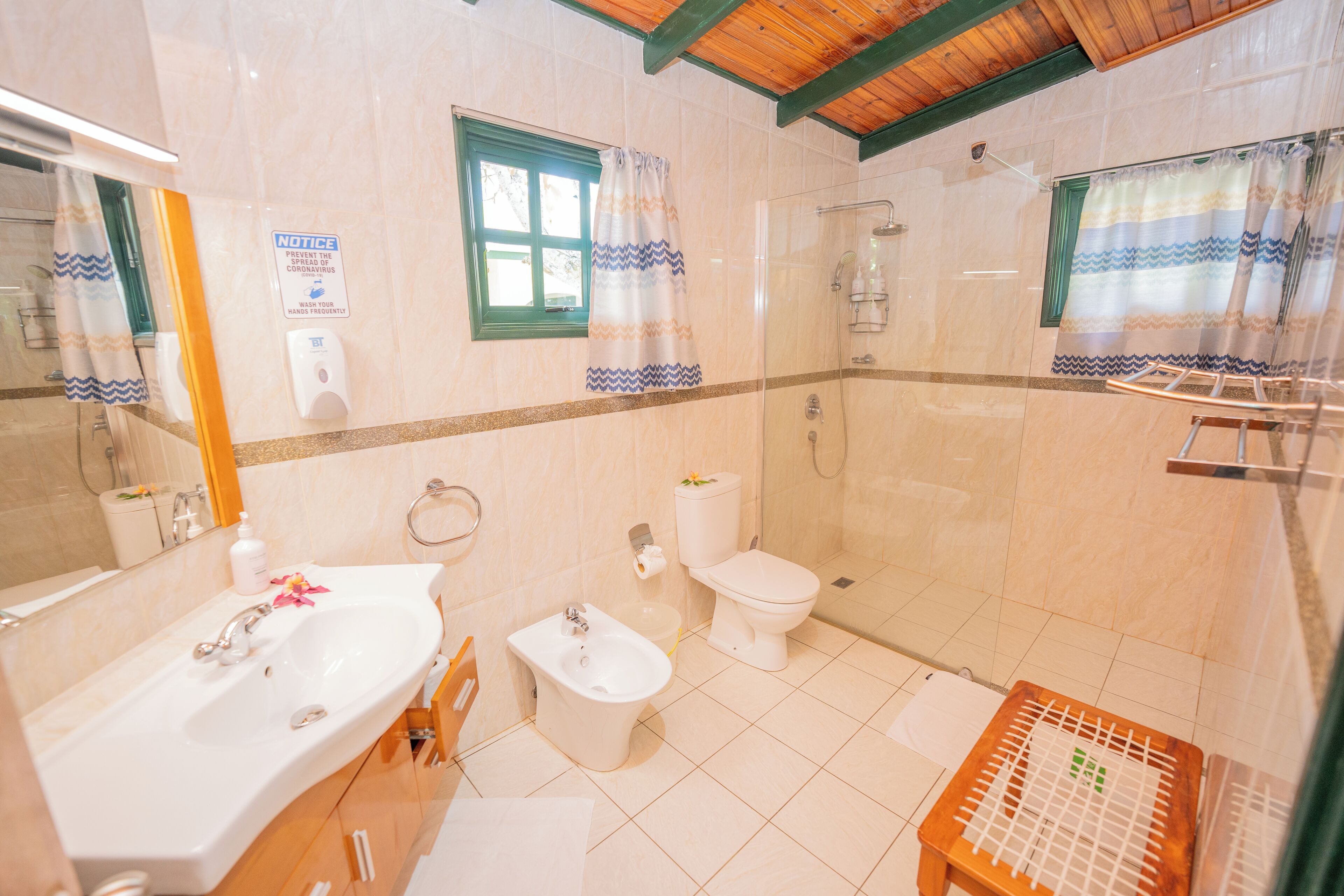 Casa de banho com lavatório, bidé, sanita e chuveiro. Há um aviso sobre lavar as mãos e uma janela com cortina.