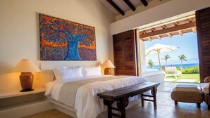 Ein Schlafzimmer mit einem Bett, Nachttischlampen, einer Bank und Blick auf das Meer durch eine offene Tür.