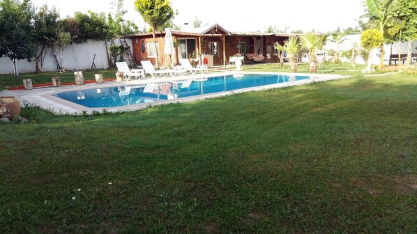 A swimming pool surrounded by a grassy lawn, with a wooden house in the background.