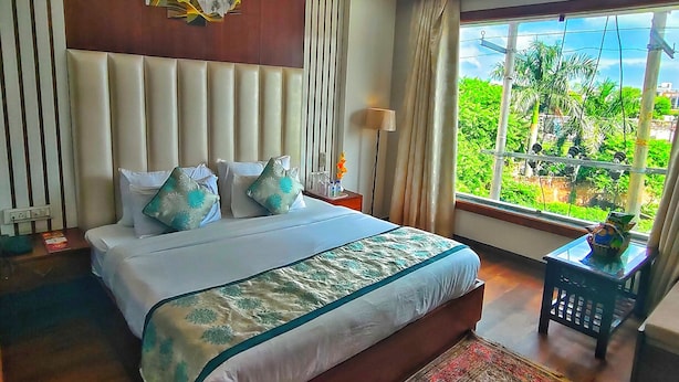 A bedroom with a large bed, a ceiling fan, a view of greenery outside, and a decorative wall piece above the bed. - HOTEL The Rich Grand (Agra)