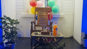 Une petite pièce décorée d’une banderole d’anniversaire, avec une chaise en bois, une petite table sur laquelle se trouve un gâteau, et des plantes en pot.