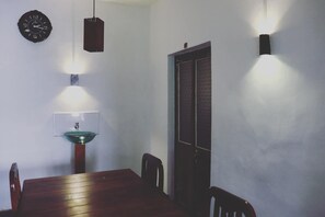 A dining area with a wooden table, chairs, a wall-mounted clock, a pendant light, and a wall-mounted light fixture. - Zion White House (Nuwara Eliya)