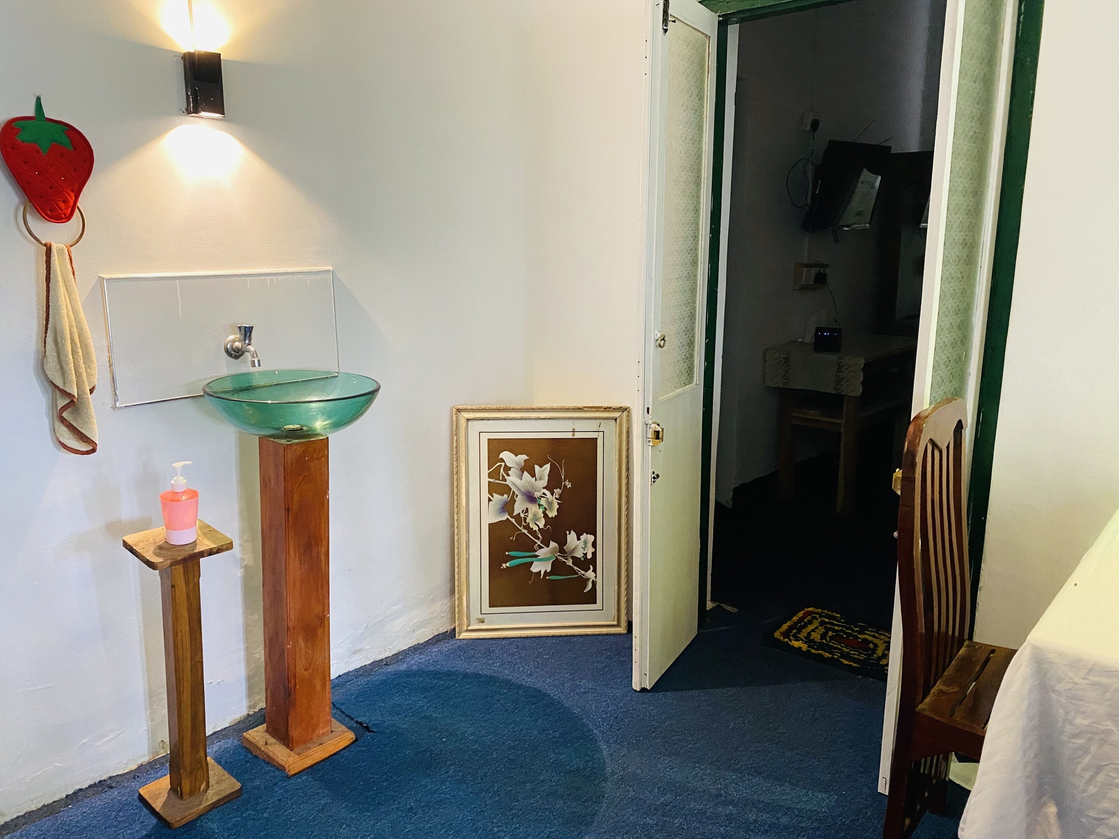 A bathroom with a glass sink, a wooden stool with a bottle, a framed picture, and a door leading to another room.