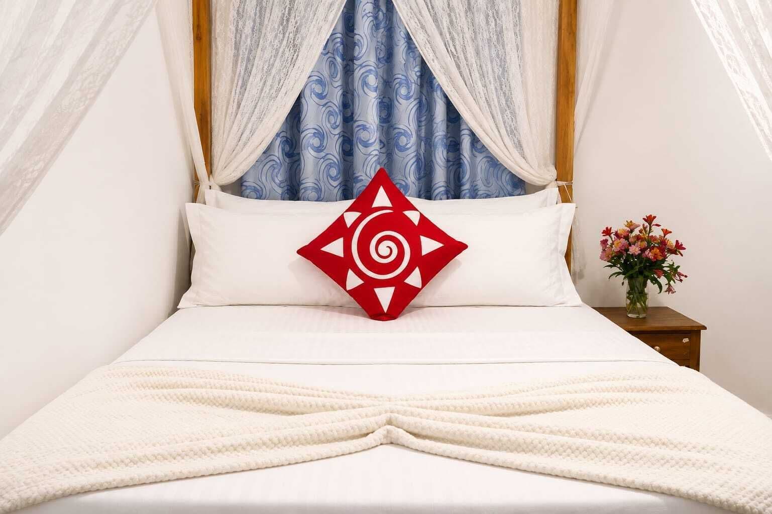A bed with a white bedspread, a red pillow with a geometric design, and a vase with flowers on a bedside table.