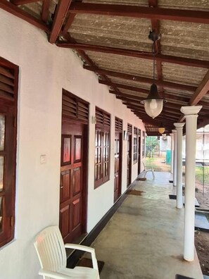 A covered corridor with wooden doors and columns, a white plastic chair, and a hanging light fixture. - Katagamuwa Rest  (Kataragama)