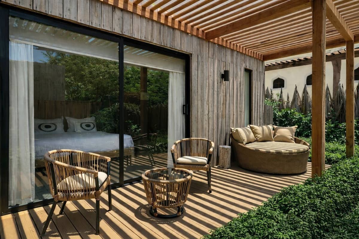 Un espacio exterior moderno con terraza de madera, puerta corrediza de vidrio que da a un dormitorio, mobiliario de mimbre y una zona de estar circular.