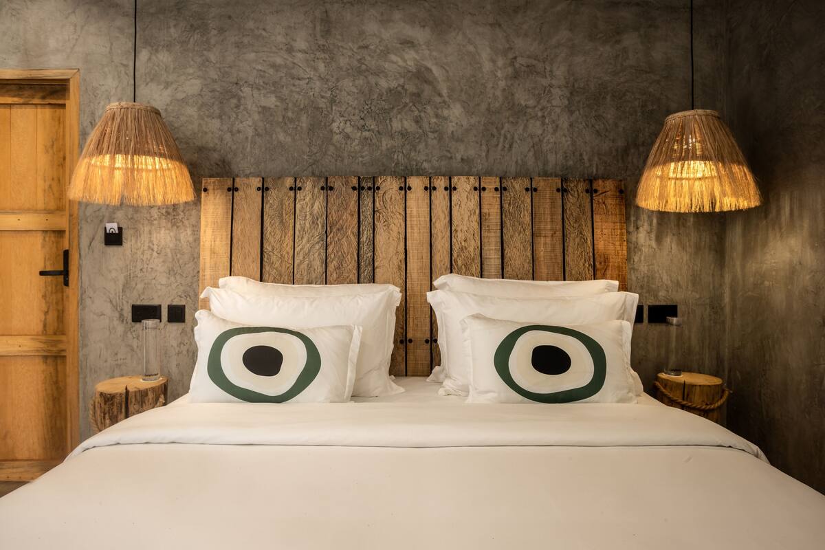 Una cama con ropa de cama blanca, almohadas verdes con forma de ojos, un cabecero de madera y dos lámparas colgantes.