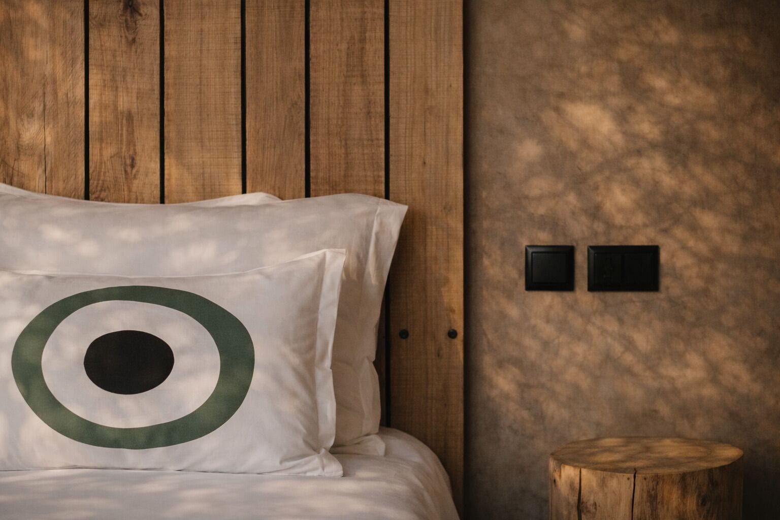 A bed with a pillow featuring a green and white design, a wooden headboard, a small wooden stool, and two wall-mounted switches.