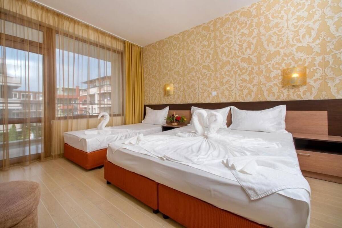 Habitación de hotel con dos camas, un ventanal grande con cortinas, una pared estampada y una pequeña mesa con un florero.