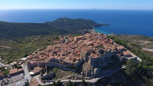 Une vue aérienne d’une ville historique, avec un imposant pont de pierre et des remparts, le tout entouré d’une végétation luxuriante et bordé par la mer.