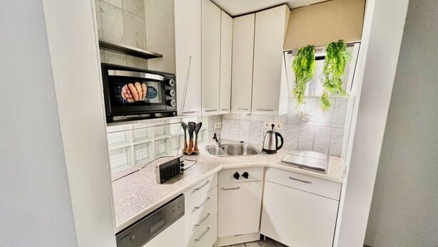 Cucina compatta con mobili bianchi, microonde, lavello e finestra con piante pensili.