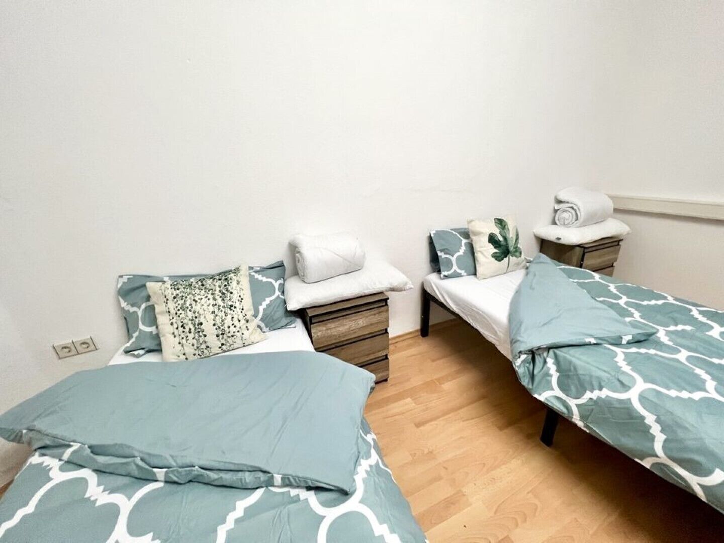 A small, tidy room with two single beds, each with a blue patterned bedspread and pillows. There are wooden nightstands with a white lamp and a rolled towel on one.