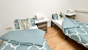 Una camera piccola e ordinata con due letti singoli, ciascuno con una coperta e dei cuscini a motivi blu. Si vedono due comodini di legno, uno con una lampada bianca e un asciugamano arrotolato.