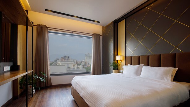 A hotel room with a large bed, a wooden floor, a window with curtains, a plant, and a city view. - Sangam City Hotel (Kathmandu)