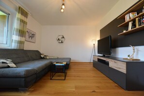 Un salon moderne avec un canapé gris, une table basse en bois, un téléviseur posé sur un meuble noir et une bibliothèque remplie de livres et d’objets décoratifs.