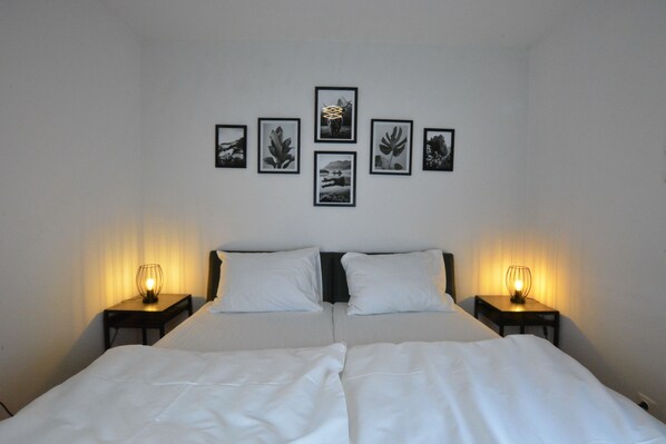 Un lit double avec du linge de lit blanc, deux tables de chevet avec des lampes et cinq tableaux encadrés accrochés au mur.