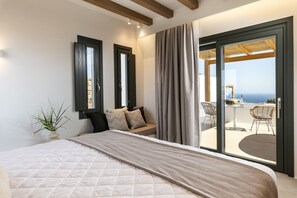 A bedroom with a large bed, a sofa, and a sliding glass door leading to an outdoor seating area with a view of the sea.
